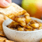 Close-up of apple pie dip with cinnamon sugar chips, featuring a chip scooping warm, spiced apple filling from a ceramic bowl.