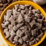 A bowl filled with brownie batter muddy buddies, coated in powdered sugar and served in a yellow dish.