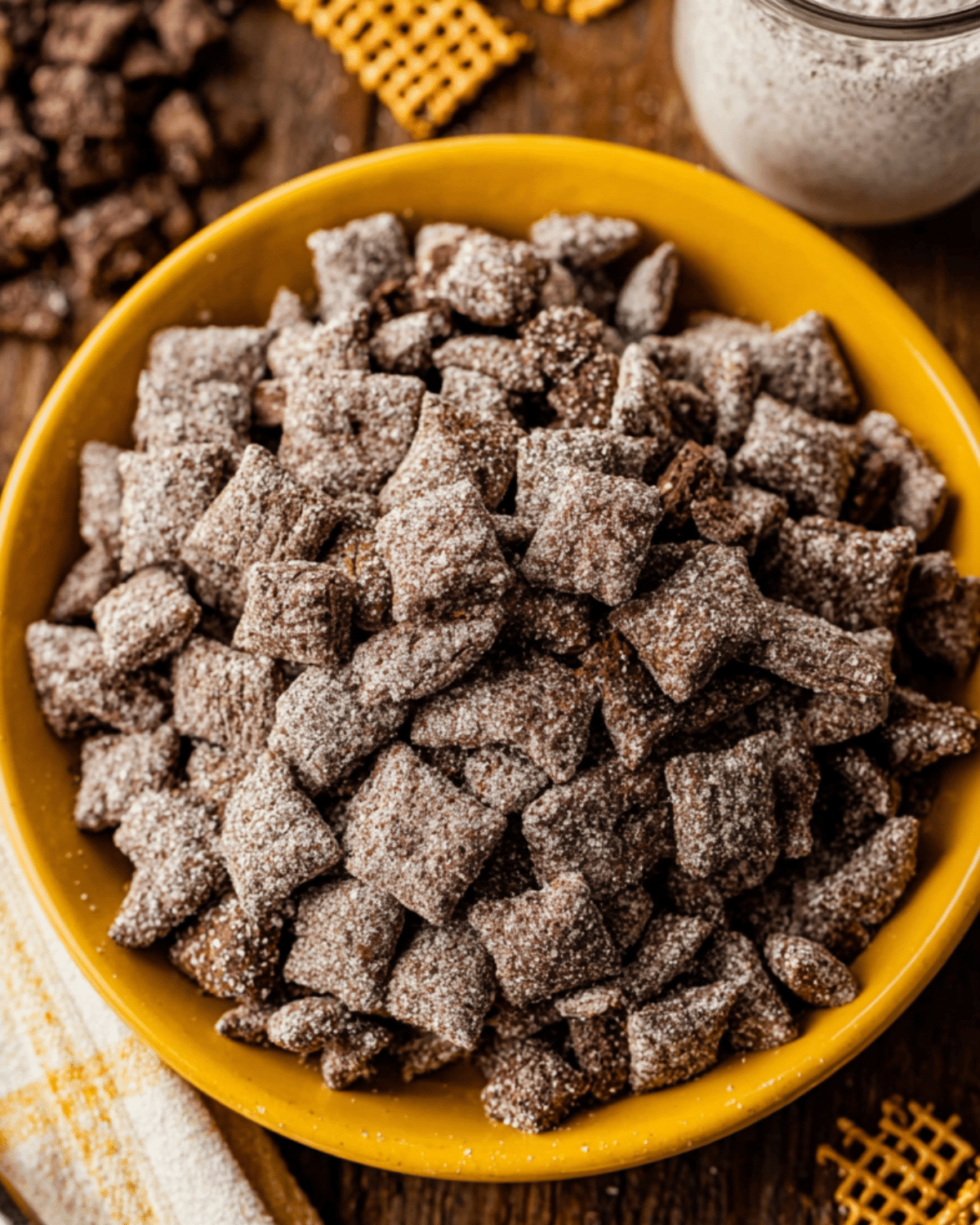 A bowl filled with brownie batter muddy buddies, coated in powdered sugar and served in a yellow dish.