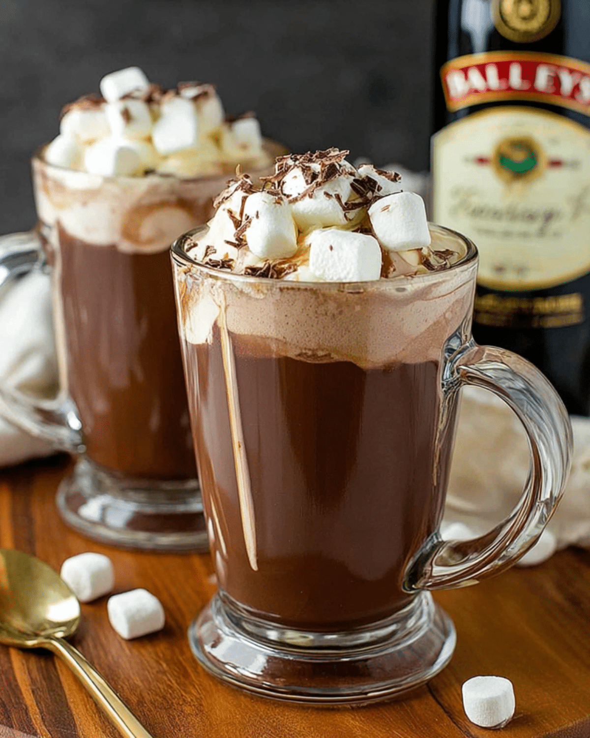 Two glass mugs of Baileys Hot Chocolate topped with whipped cream, mini marshmallows, and chocolate shavings on a wooden surface.
