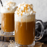 Two glass mugs of Butterbeer topped with whipped cream and drizzled caramel, set on a rustic wooden surface.
