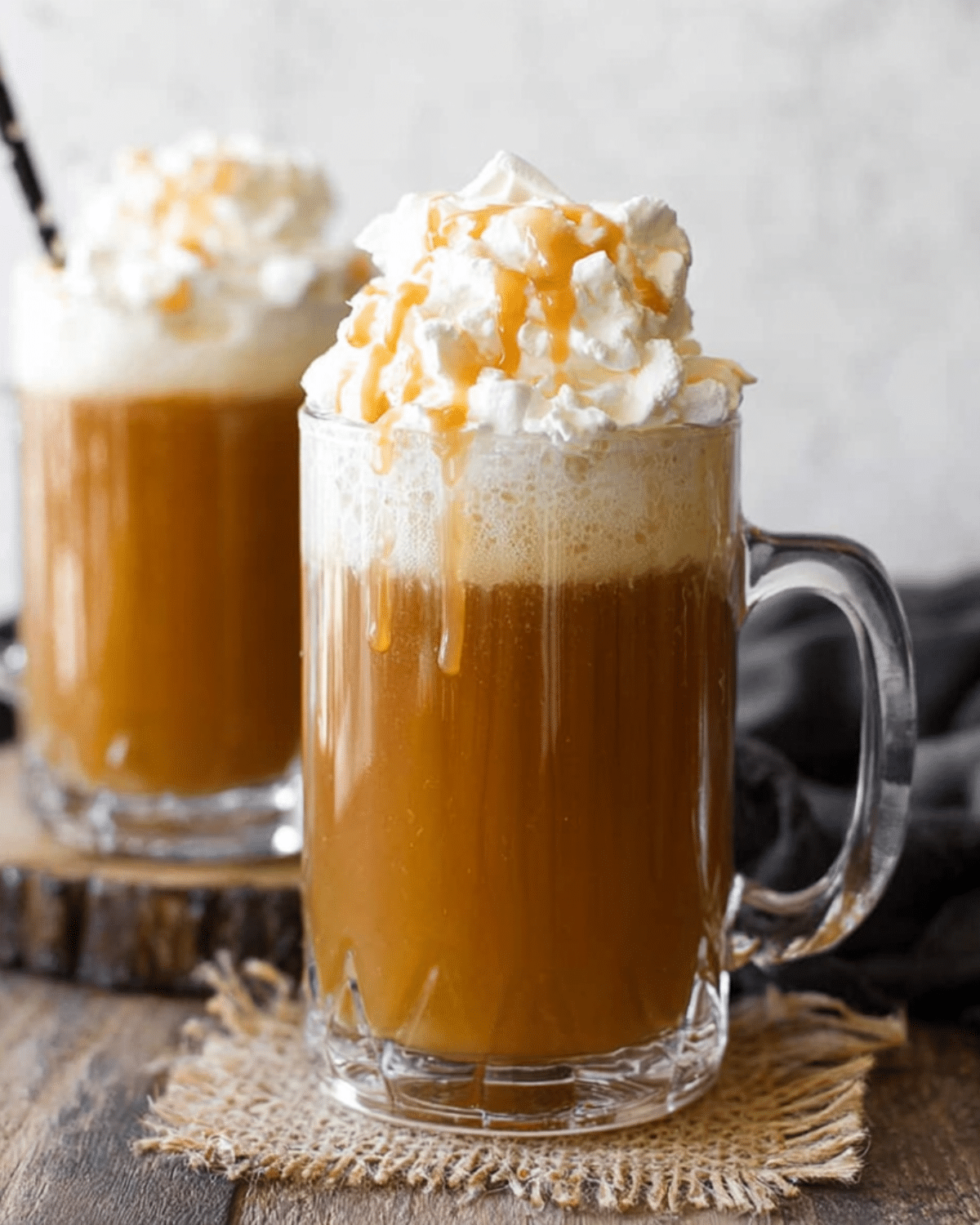 Two glass mugs of Butterbeer topped with whipped cream and drizzled caramel, set on a rustic wooden surface.