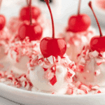 White chocolate-covered CHRISTMAS CHERRY BOMBS topped with crushed peppermint and bright red cherries on a white plate.