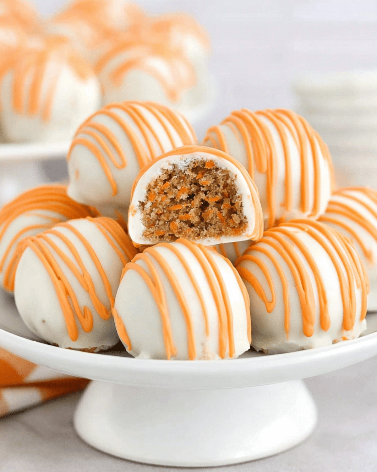 Carrot cake cheesecake bites coated in white chocolate and drizzled with orange icing, displayed on a white cake stand.