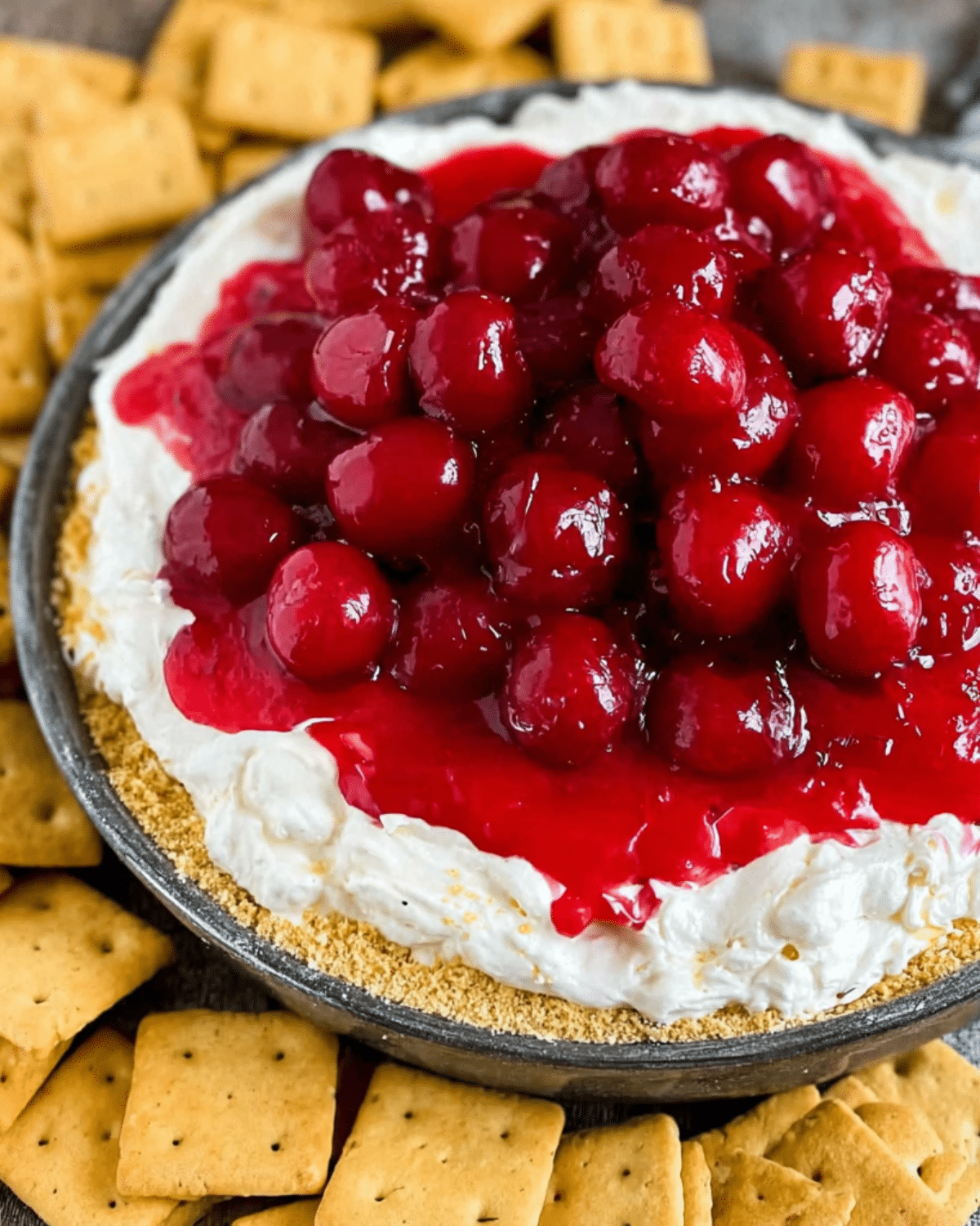 Close-up of cherry cheesecake dip topped with glossy cherry pie filling, surrounded by square graham crackers.