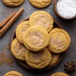 A wooden plate stacked with Chewy Pumpkin Cookies, dusted with sugar, surrounded by cinnamon sticks and spices.
