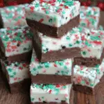 Stacked squares of Christmas fudge with a chocolate base and white top layer, decorated with red, green, and white holiday sprinkles.