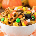 A white bowl filled with Easy Halloween Snack Mix featuring candy corn, pretzels, cereal, chocolates, and a fake spider.