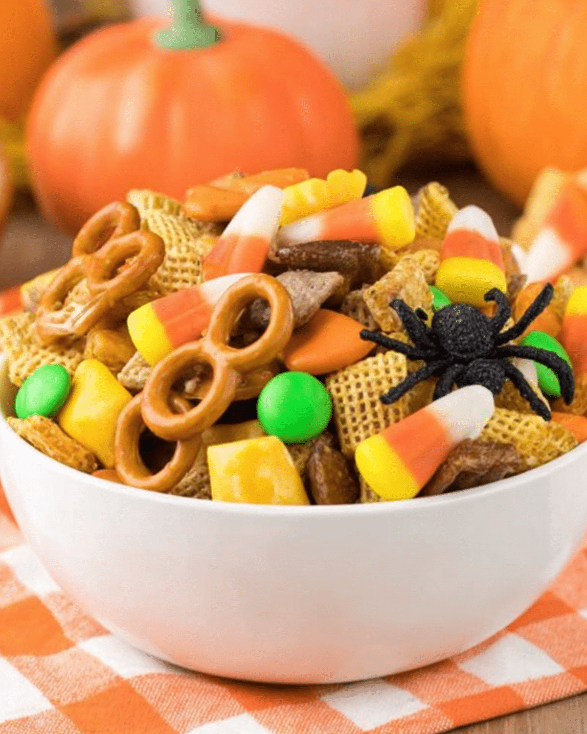 A white bowl filled with Easy Halloween Snack Mix featuring candy corn, pretzels, cereal, chocolates, and a fake spider.
