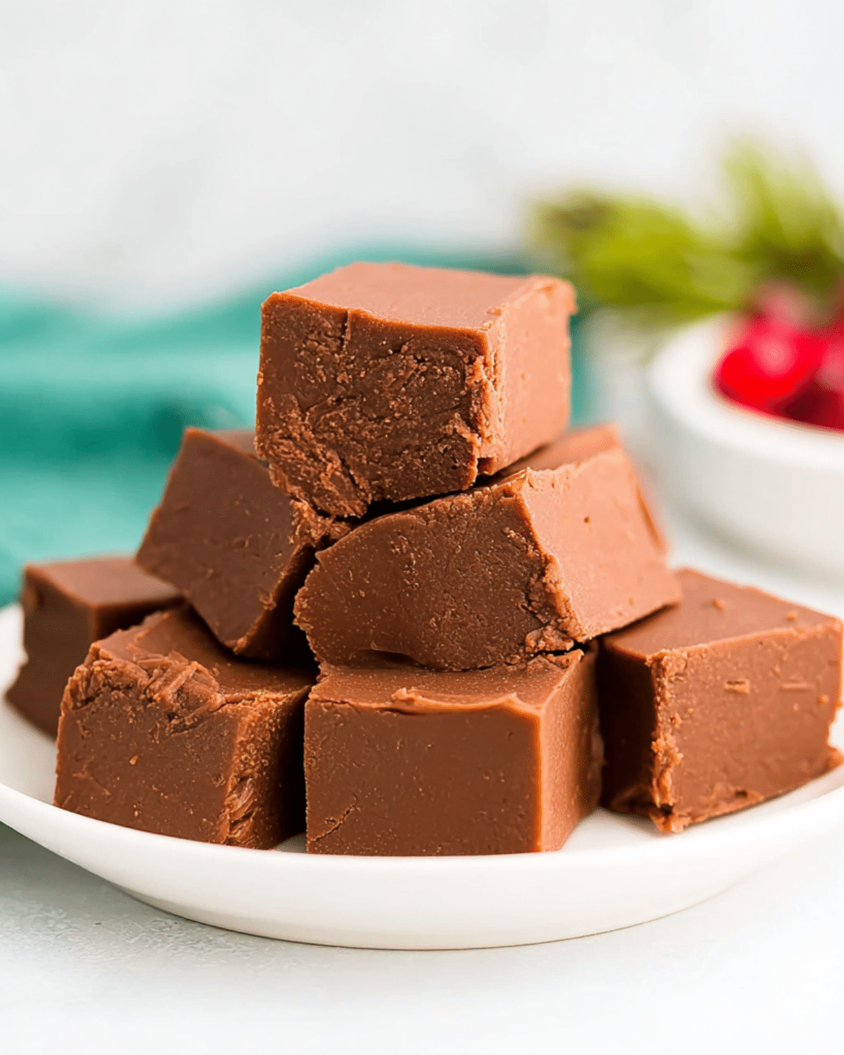 Stack of rich, creamy fantasy fudge squares on a white plate, with a festive background of cranberries and greenery.