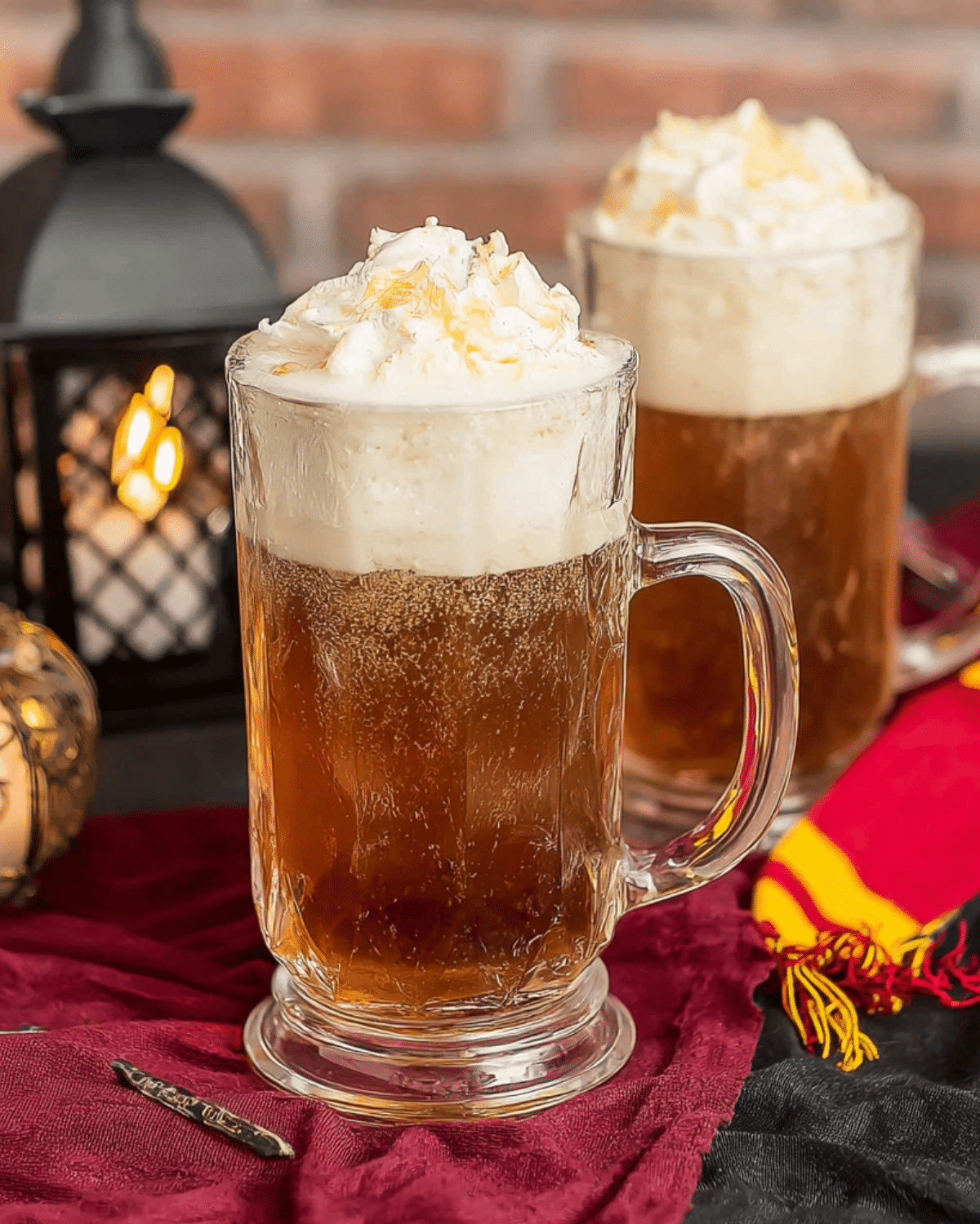 Glass mugs filled with frothy Harry Potter Butterbeer topped with whipped cream and butterscotch drizzle, set on a burgundy cloth with a lantern in the background.