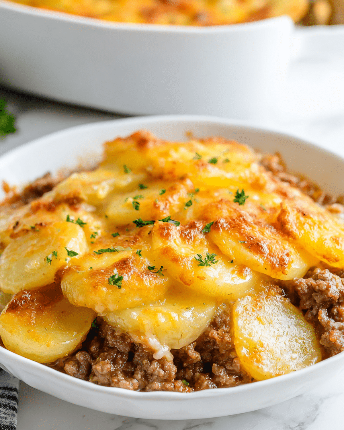 Hamburger & potato casserole topped with golden baked sliced potatoes and melted cheese in a white bowl.