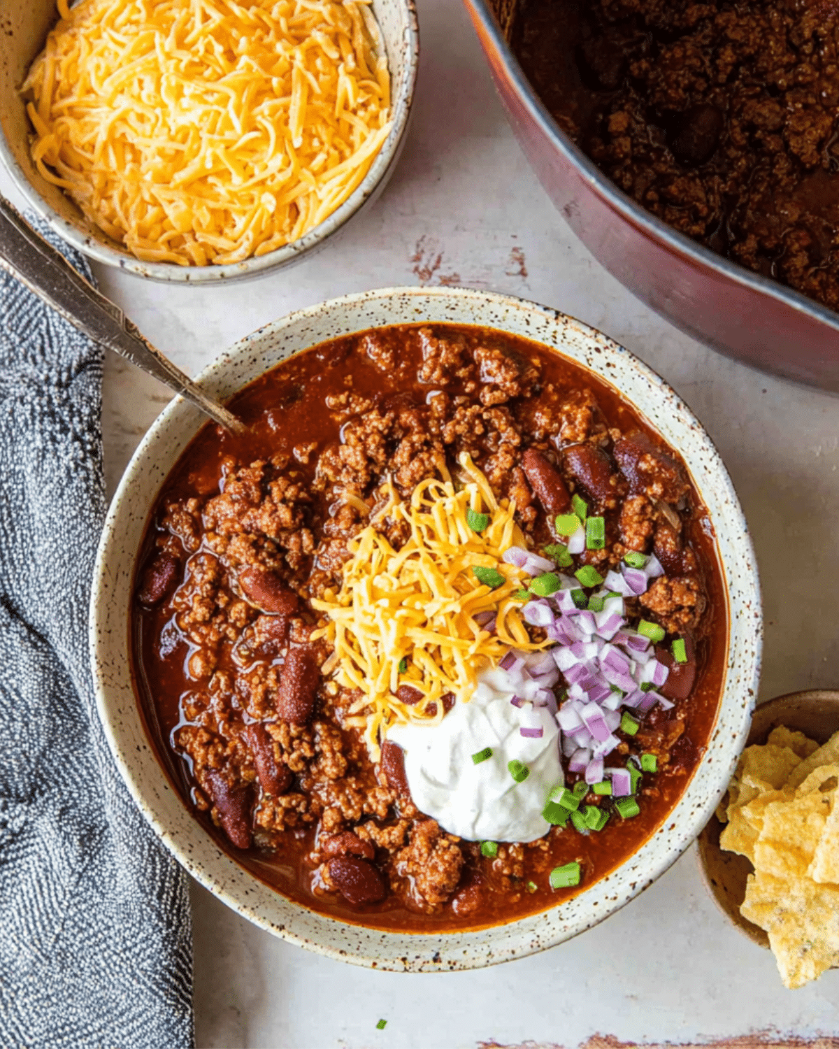 Homemade Ground Beef Chili in a bowl topped with shredded cheese, sour cream, chopped red onions, and green onions, served with tortilla chips on the side.
