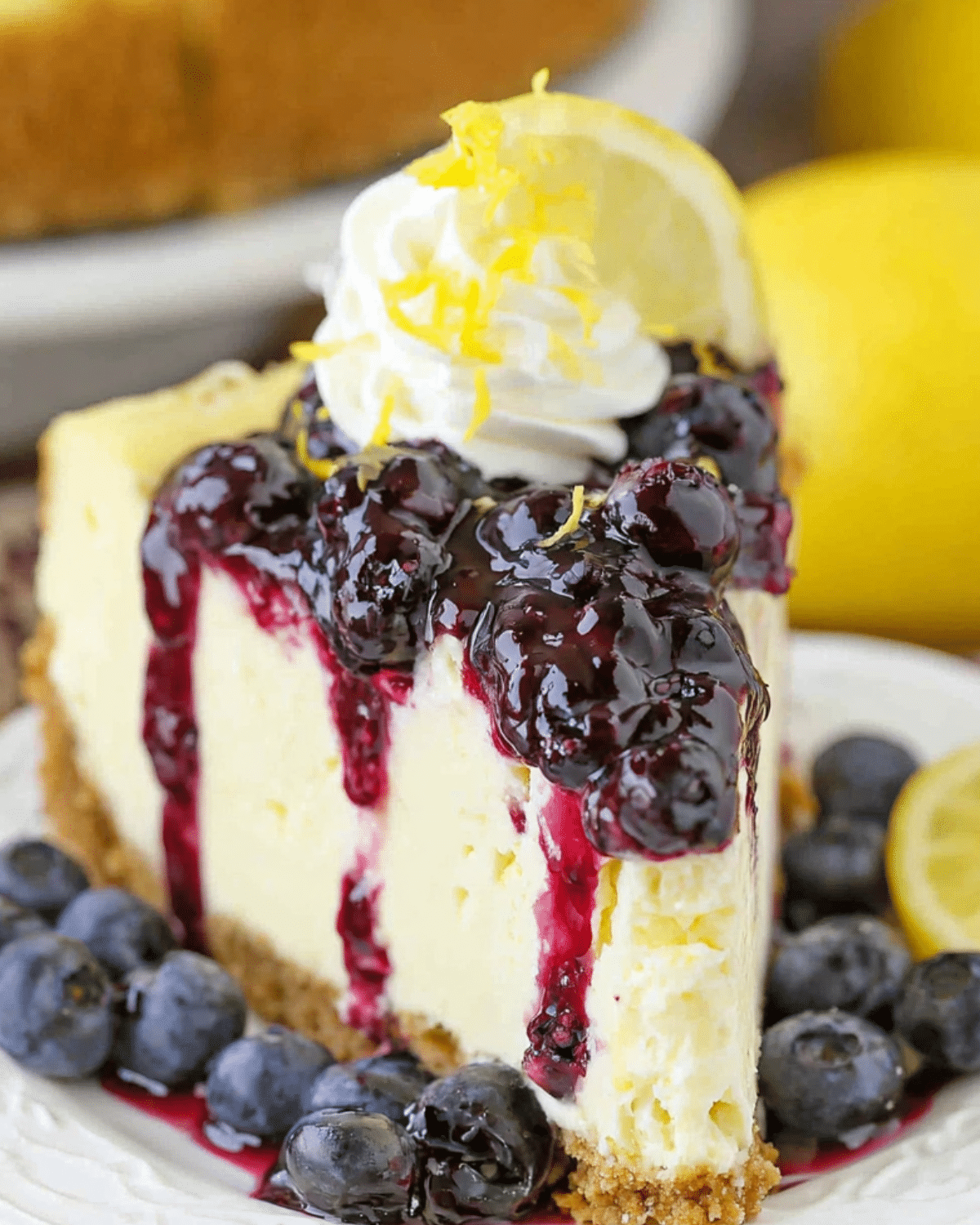 Slice of lemon blueberry cheesecake topped with blueberry compote, whipped cream, lemon zest, and fresh blueberries.