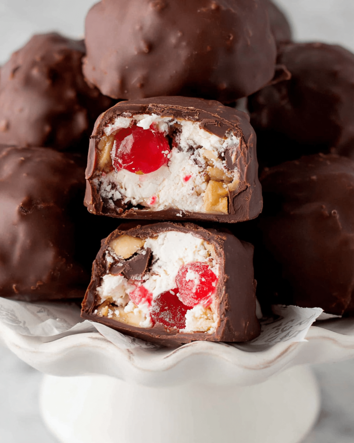 Close-up of Martha Washington Candy filled with cherries, coconut, and pecans, coated in chocolate and stacked on a white pedestal plate.