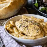 A bowl of creamy Olive Garden chicken served over penne pasta with herbs, accompanied by fresh bread and salad in the background.