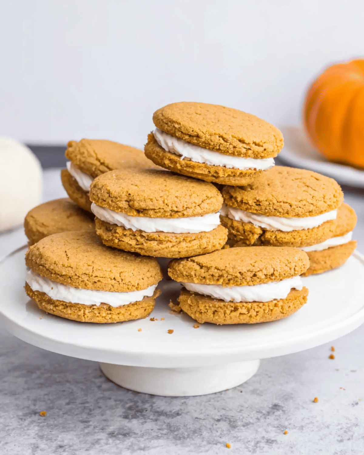 A stack of Pumpkin Pie Cookie Sandwiches with creamy white filling on a white cake stand.