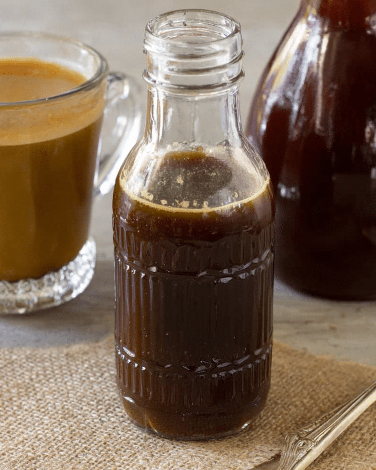 A glass bottle filled with homemade Starbucks Pumpkin Spice Coffee Syrup on a burlap cloth, with a cup of pumpkin spice coffee and another syrup bottle in the background.