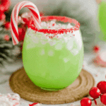 A festive glass of The Grinch Punch with a red sugar rim, crushed peppermint, and a candy cane garnish.