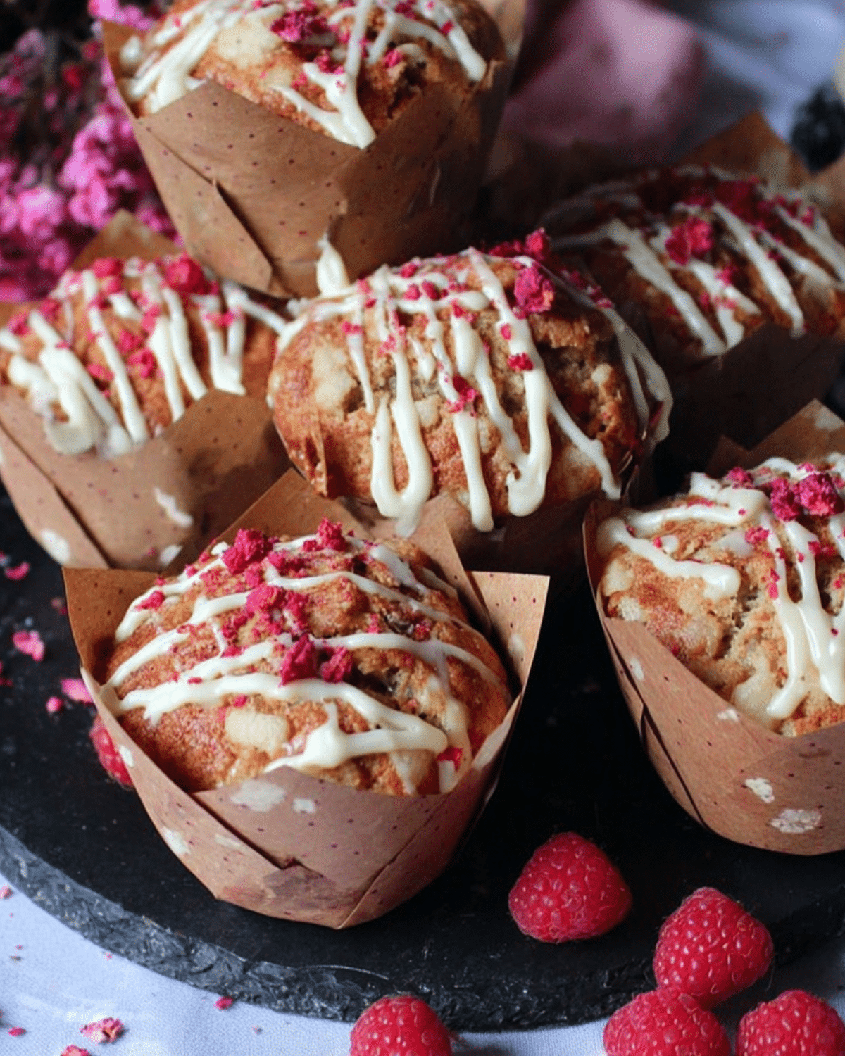 White chocolate and raspberry muffins drizzled with white chocolate and topped with crushed freeze-dried raspberries, surrounded by fresh raspberries.