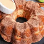 Apple cider donut cake covered in cinnamon sugar, presented on a black plate with a creamy glaze being poured over the top.