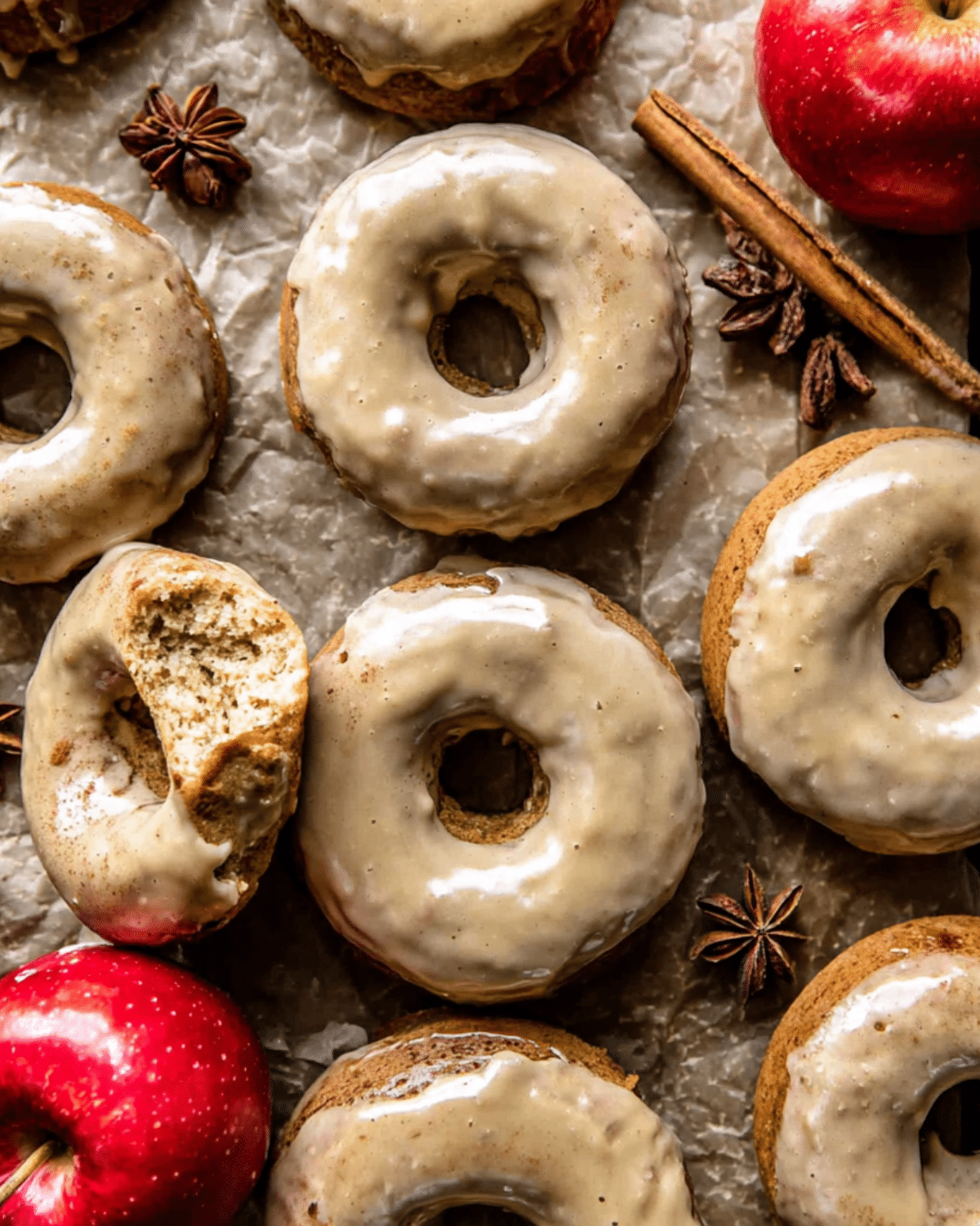 A batch of baked apple cider doughnuts topped with a shiny cinnamon maple glaze, surrounded by fresh apples, cinnamon sticks, and star anise.