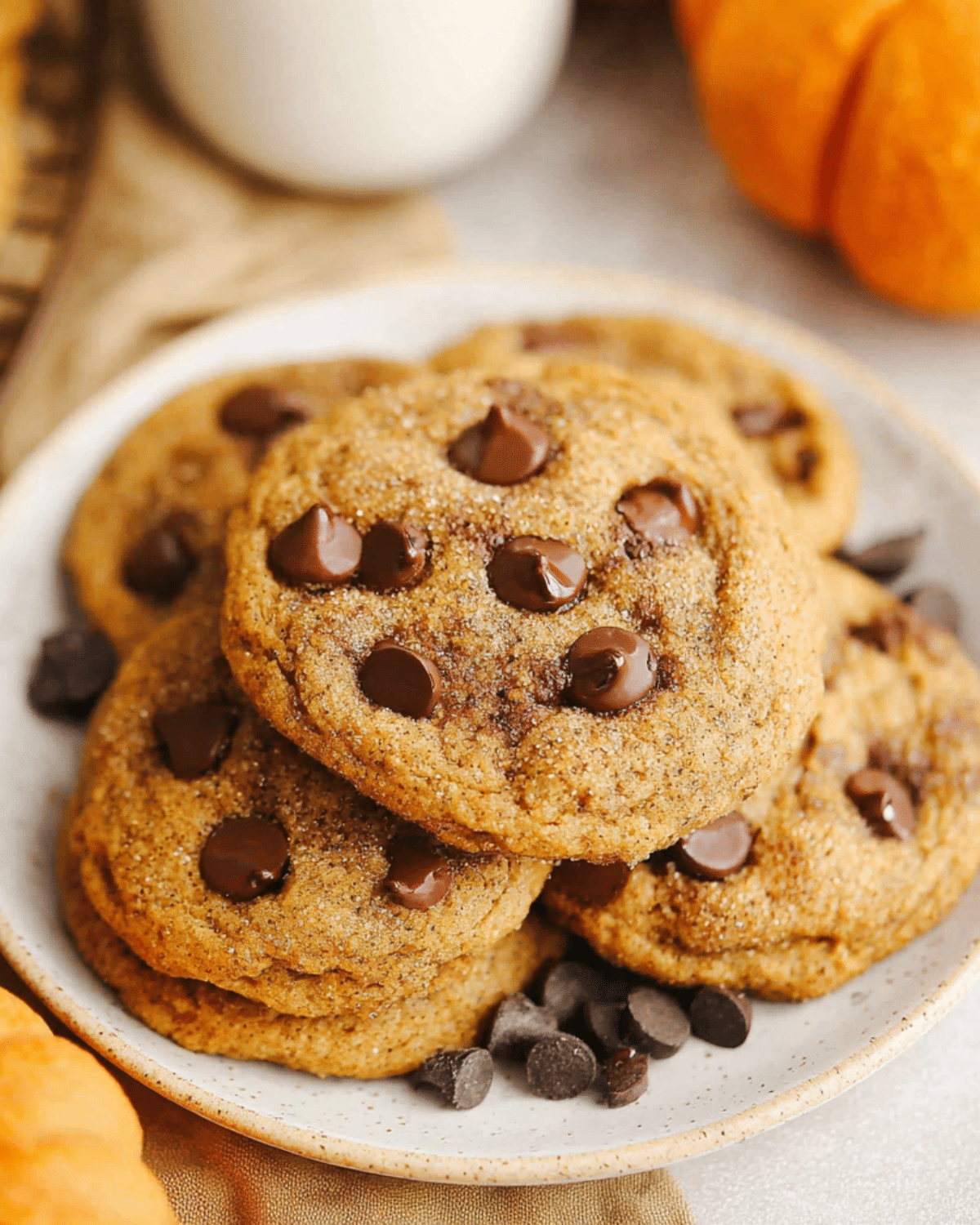A plate of brown butter pumpkin chocolate chip cookies topped with melted chocolate chips, surrounded by chocolate pieces and pumpkins.