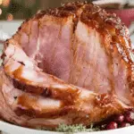 Brown sugar glazed ham with a caramelized crust, partially sliced and served on a white platter with herbs and cranberries.