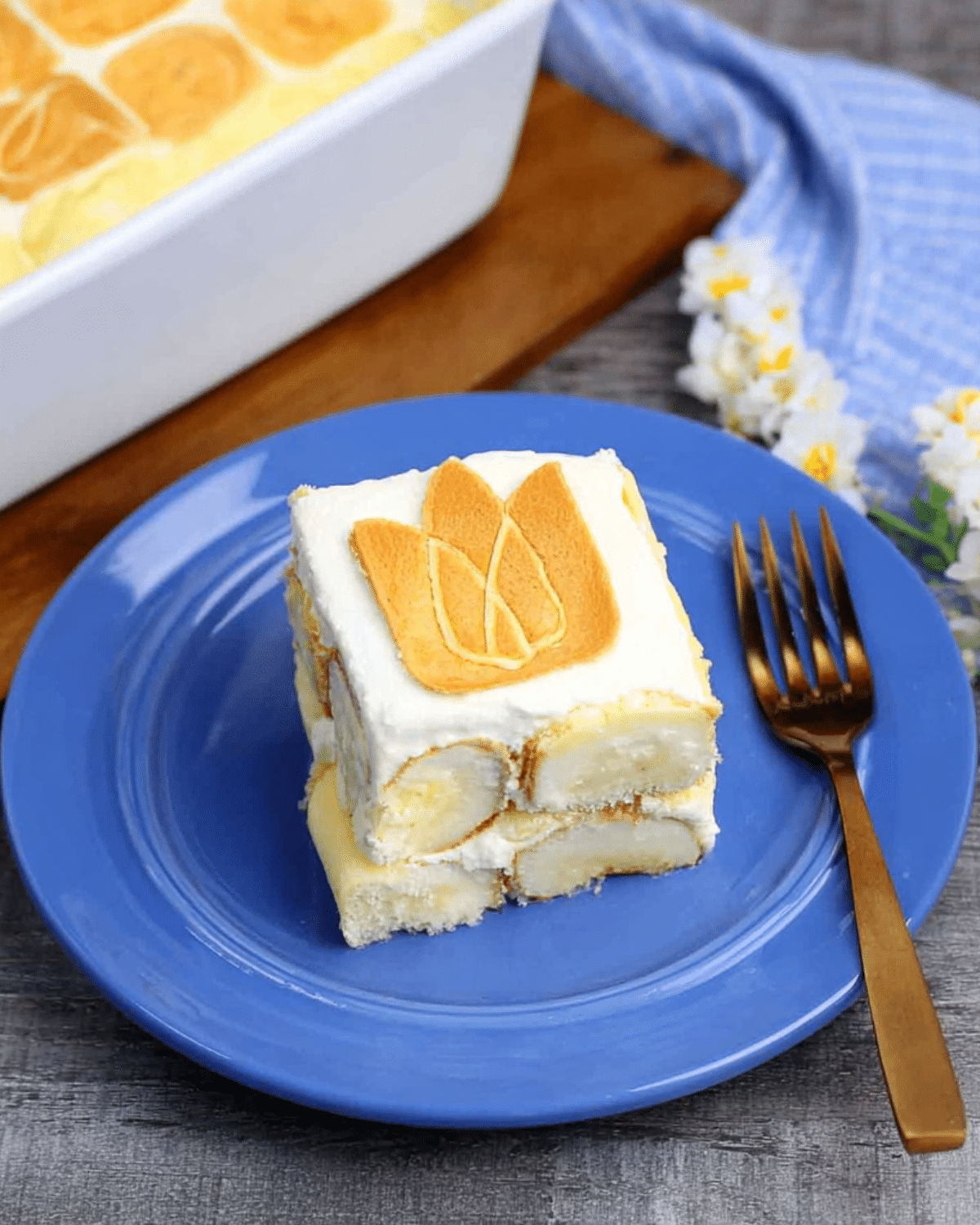 A square slice of Chessman banana pudding served on a blue plate, topped with a signature Chessman cookie and layered with banana slices, creamy pudding, and whipped topping.