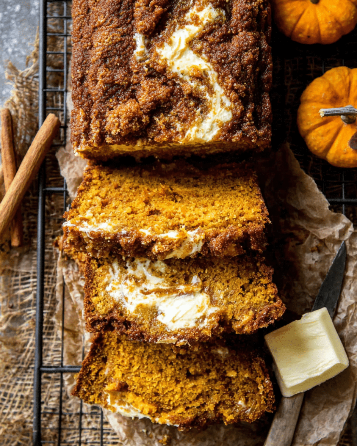 Sliced cream cheese swirled pumpkin butter bread on a wire rack, with a rustic background, butter, cinnamon sticks, and mini pumpkins.