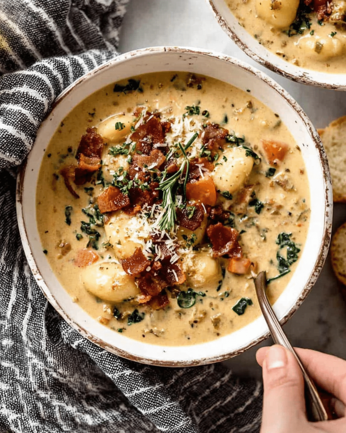 A bowl of creamy gnocchi soup topped with crispy rosemary bacon, shredded cheese, and herbs, served with a spoon on a rustic napkin.