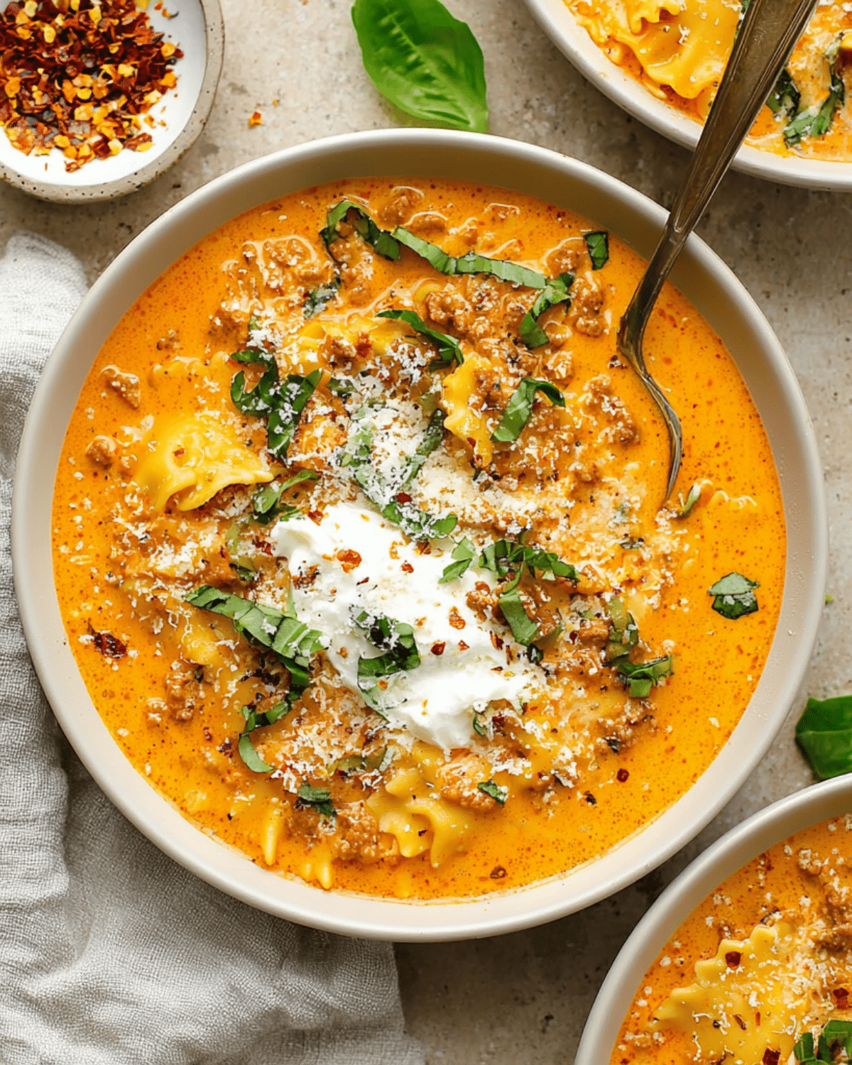 A bowl of creamy lasagna soup with ground meat, lasagna noodles, topped with ricotta, grated cheese, basil, and red pepper flakes.