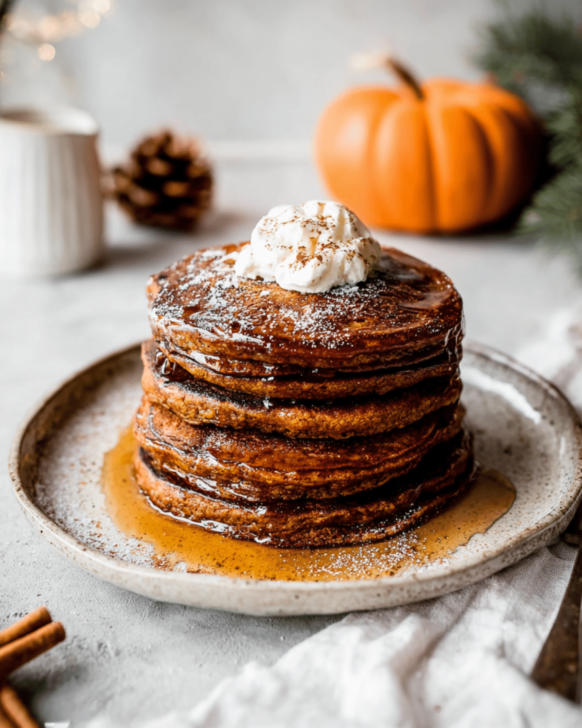 A tall stack of gingerbread pancakes topped with whipped cream and maple syrup, dusted with powdered sugar and cinnamon, with a pumpkin in the background.