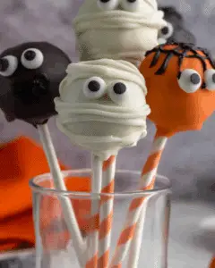 A festive assortment of Halloween cake pops decorated as mummies, monsters, and pumpkins on striped sticks in a glass jar.