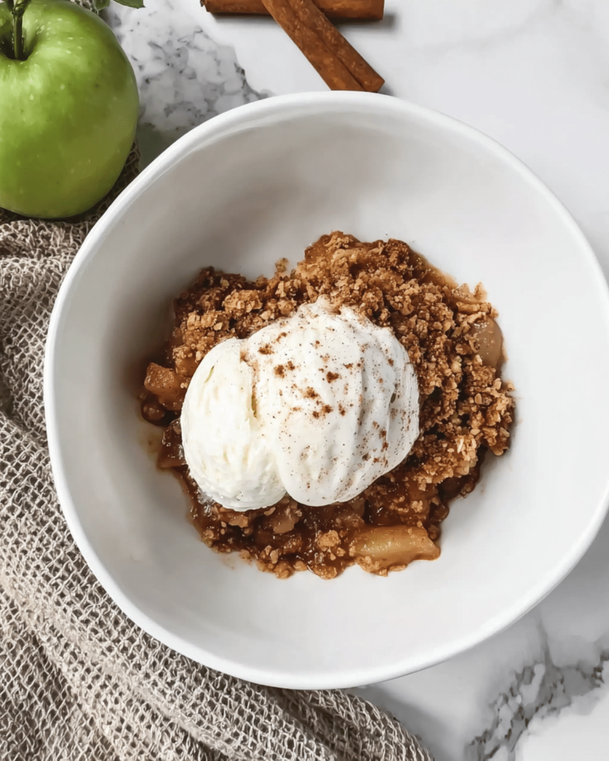 Bowl of harvest spiced apple crumble topped with a scoop of vanilla ice cream and dusted with cinnamon, served on a marble surface with a green apple and cinnamon sticks nearby.