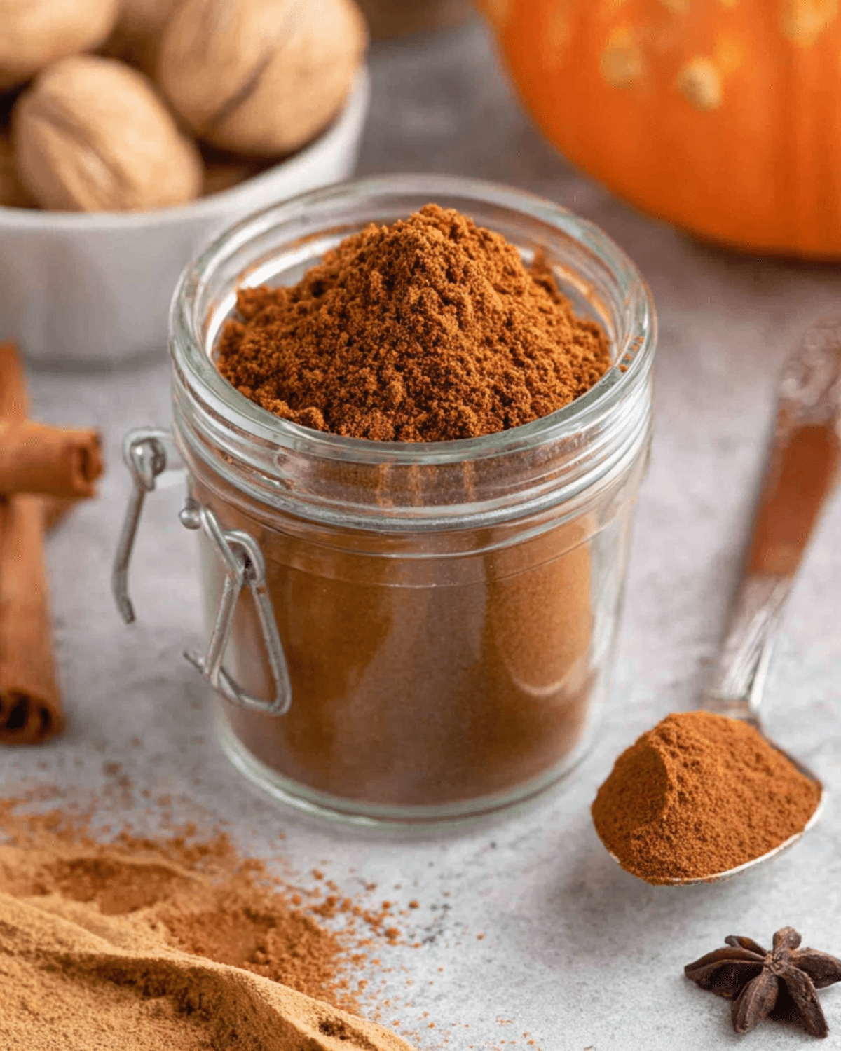 Homemade pumpkin pie spice in a glass jar with a spoonful beside it, surrounded by cinnamon sticks, whole nutmeg, and a pumpkin.