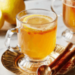 A glass mug of hot toddy cocktail garnished with a lemon slice, served on a gold-rimmed saucer with cinnamon sticks and a golden spoon.