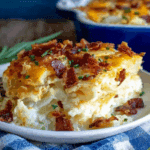 A serving of loaded scalloped potatoes topped with crispy bacon, melted cheese, and chopped chives on a white plate.