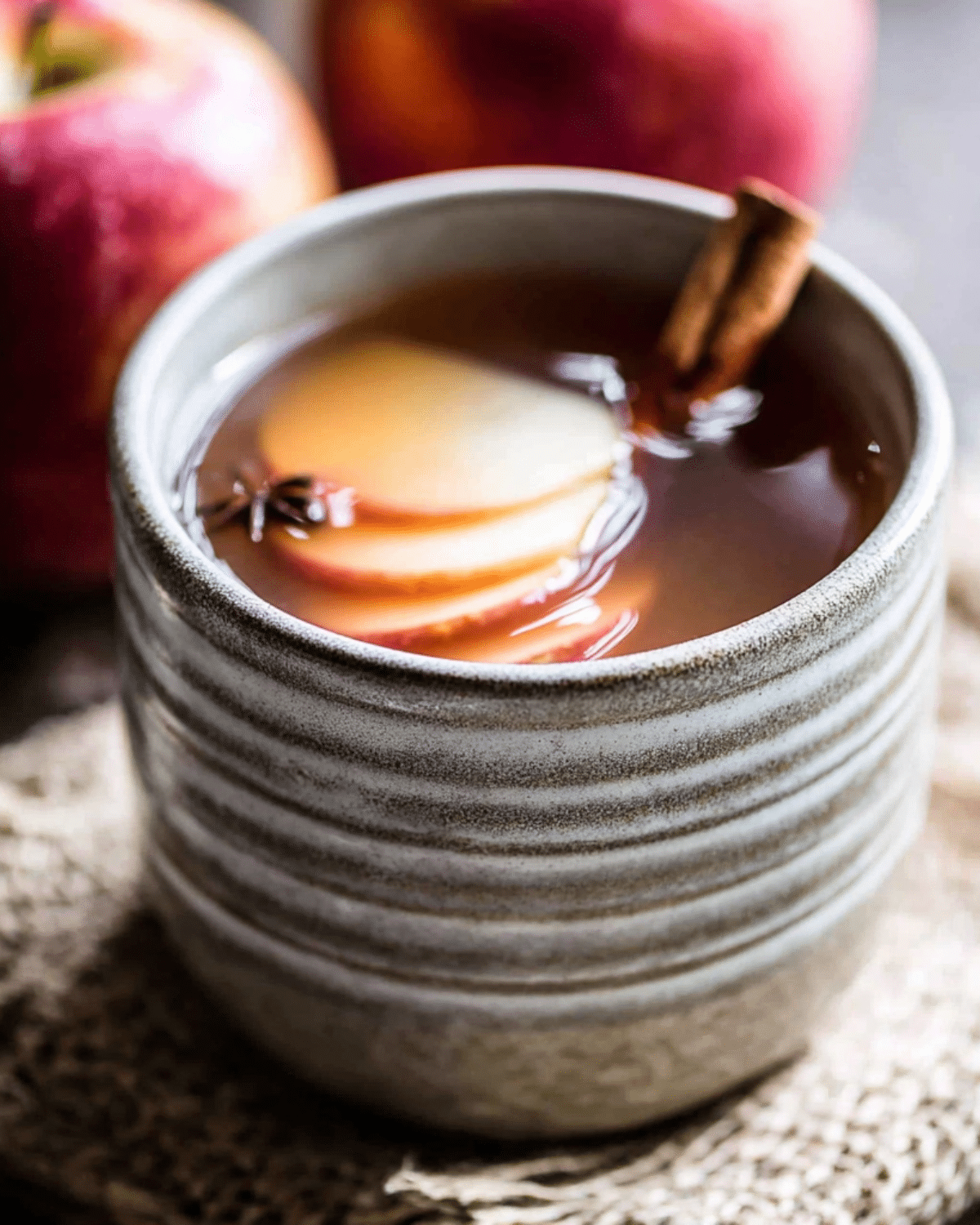 Warm maple apple cider in a rustic ceramic mug, garnished with apple slices, a cinnamon stick, and star anise.