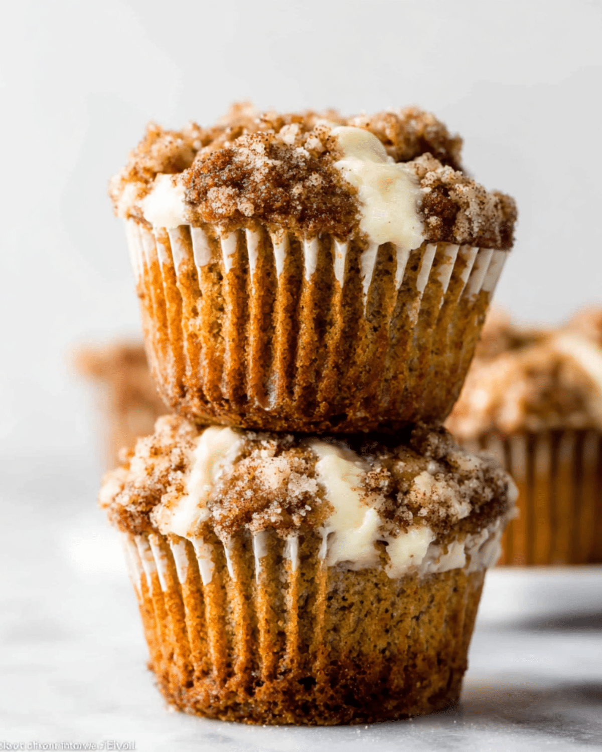 Pumpkin cream cheese muffins stacked on top of each other, topped with a cinnamon sugar crumble and filled with creamy cheesecake swirls.