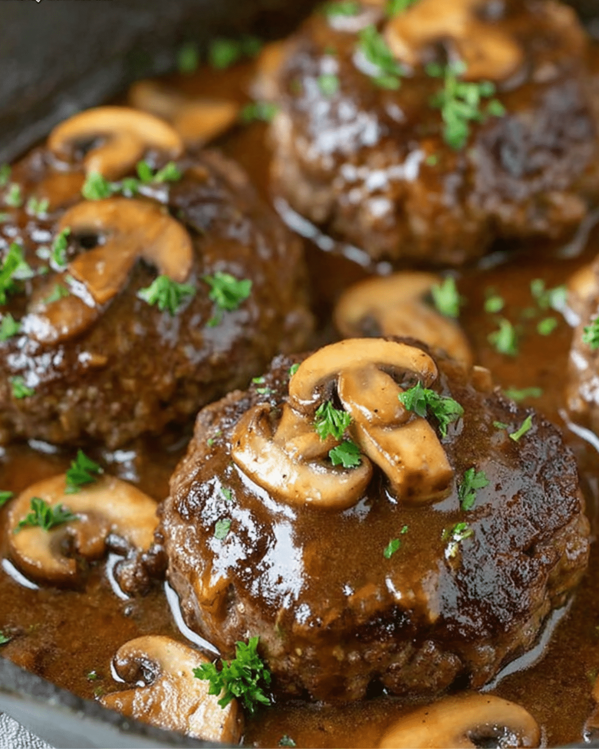 Juicy Salisbury steak patties smothered in rich brown mushroom gravy, garnished with parsley in a cast iron skillet.