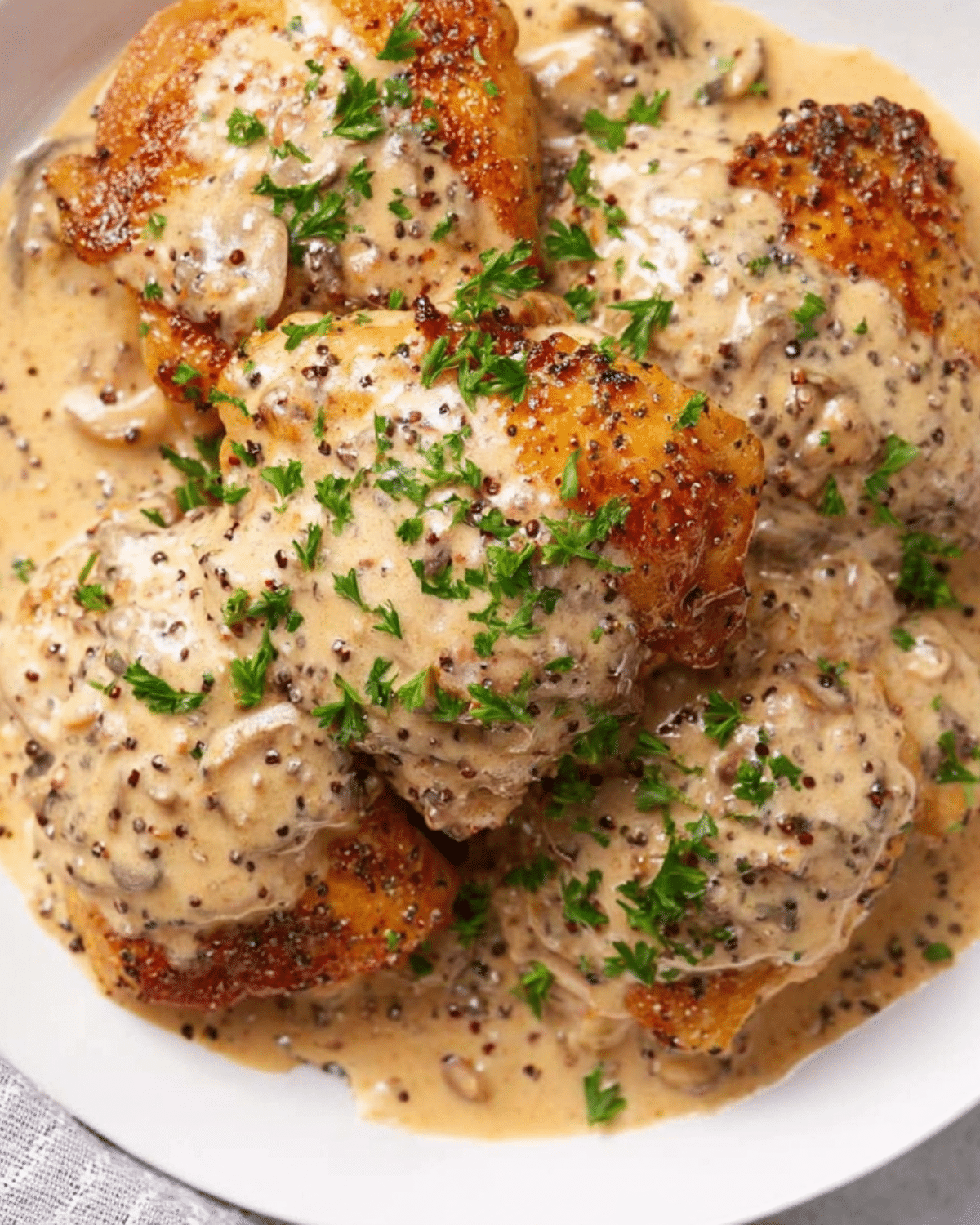 Golden seared chicken thighs covered in a creamy French wine and mustard sauce, garnished with fresh parsley on a white plate.