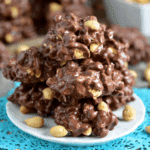 Toffee Peanut Clusters stacked on a white plate with peanuts scattered around and a blue doily underneath.