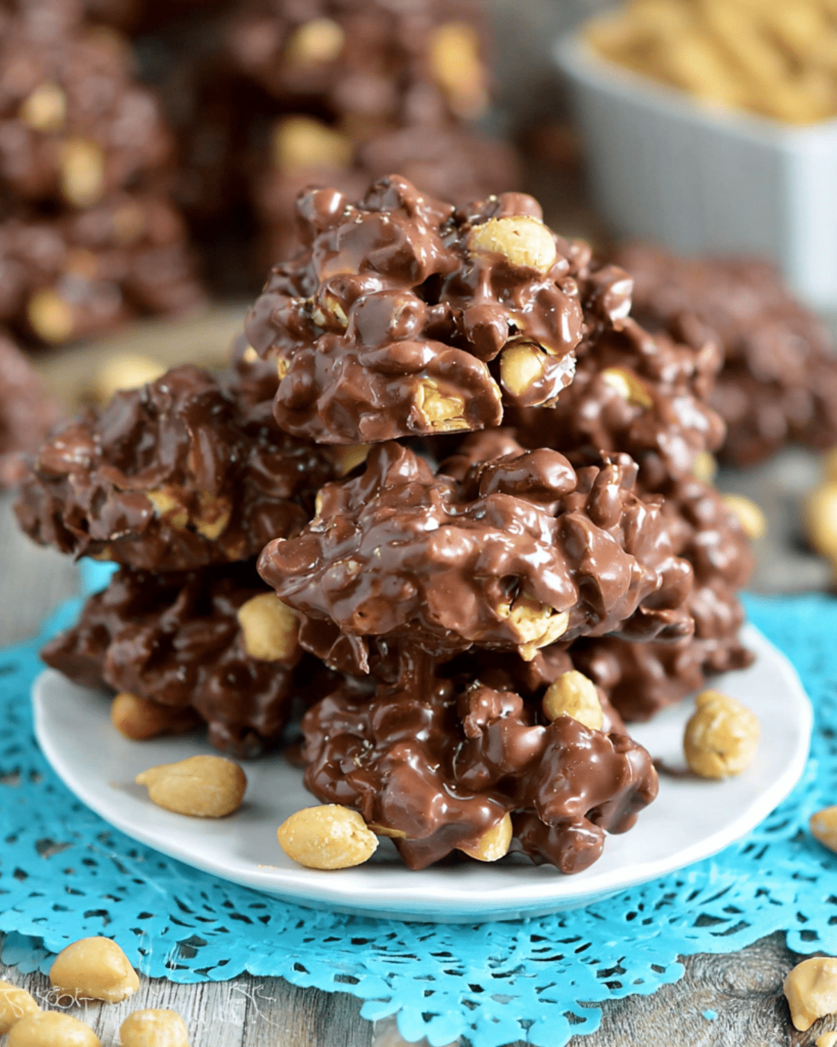 Toffee Peanut Clusters stacked on a white plate with peanuts scattered around and a blue doily underneath.