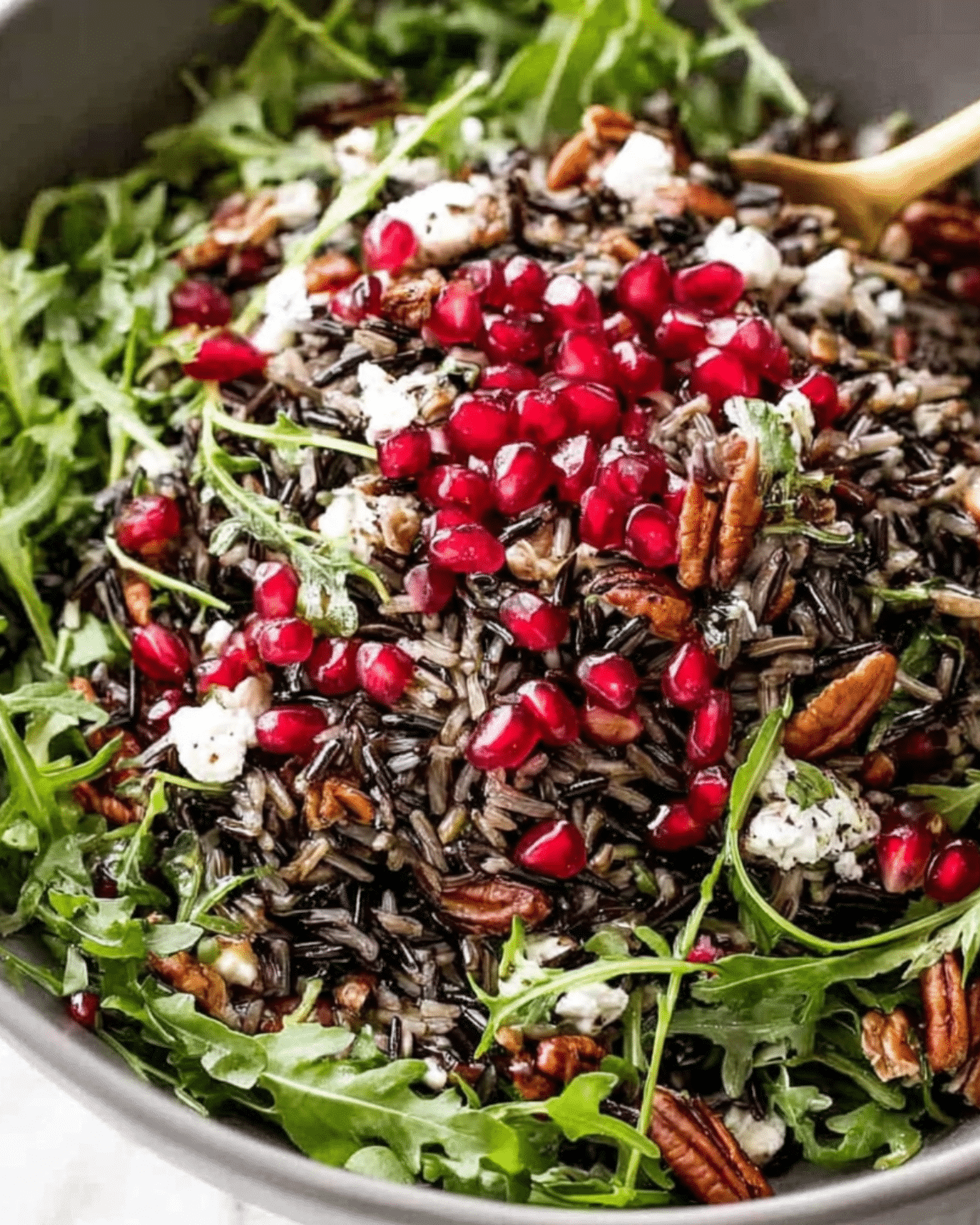 Wild rice salad with arugula, pomegranate seeds, pecans, and crumbled cheese in a large serving bowl.