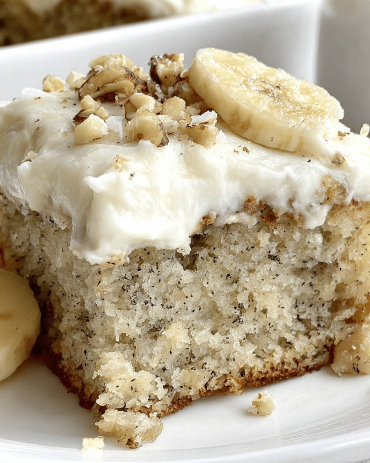 A moist slice of banana bread cake topped with cream cheese frosting, chopped walnuts, and a banana slice.