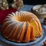 A golden boozy eggnog Bundt cake dusted with powdered sugar, served on a festive blue plate with a slice cut out.