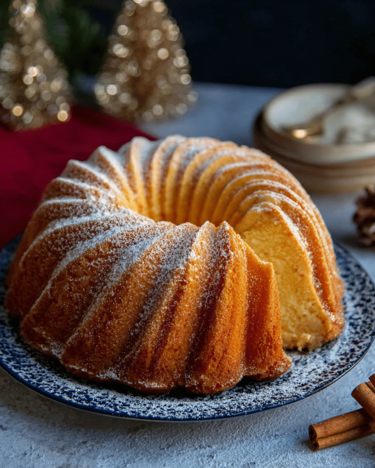 A golden boozy eggnog Bundt cake dusted with powdered sugar, served on a festive blue plate with a slice cut out.