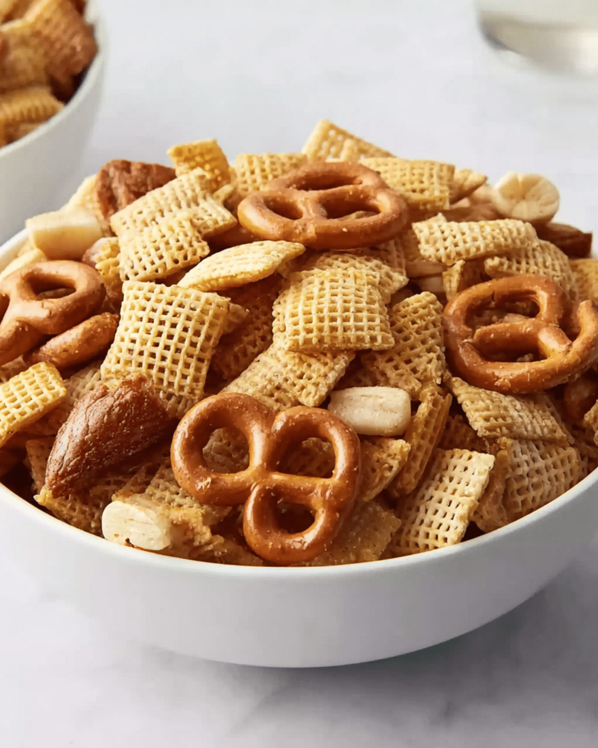 Chex party mix in a white bowl featuring cereal squares, pretzels, nuts, and seasoning.