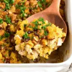 Spoonful of chicken and stuffing casserole with golden cornbread topping, cranberries, and parsley in a white baking dish.